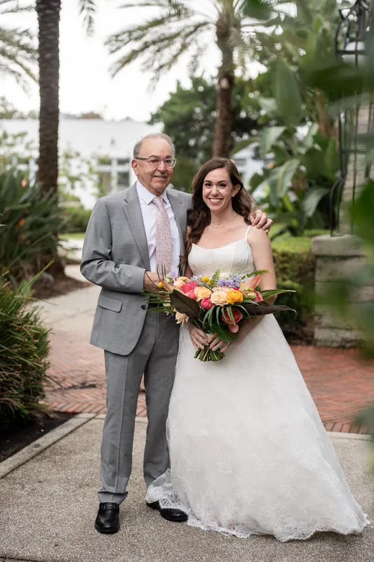 Jillian & Jason stand together at Lightner Museum's tropical courtyard, holding a vibrant bouquet with orange and red flowers.