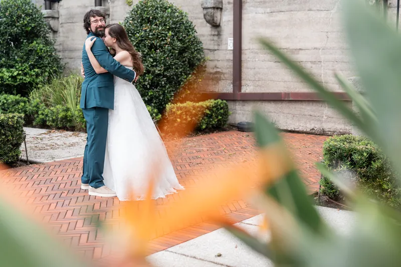 Two people embrace on a brick pathway lined with topiaries at Lightner Museum, framed by orange flowers in soft focus.