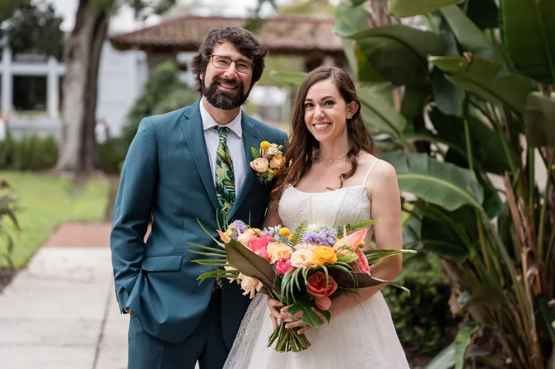A couple poses outdoors at Lightner Museum holding a colorful bouquet, both smiling in wedding attire against lush greenery.