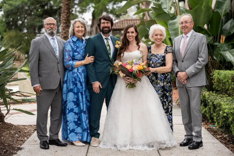 Six family members pose together outdoors, with someone in a white gown holding a bouquet amid palm trees at Lightner Museum, St. Augustine.