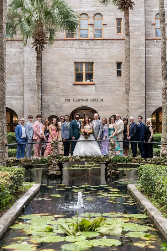 Large wedding group stands before a fountain in the courtyard of a historic stone building with palm trees at Lightner Museum, St. Augustine.
