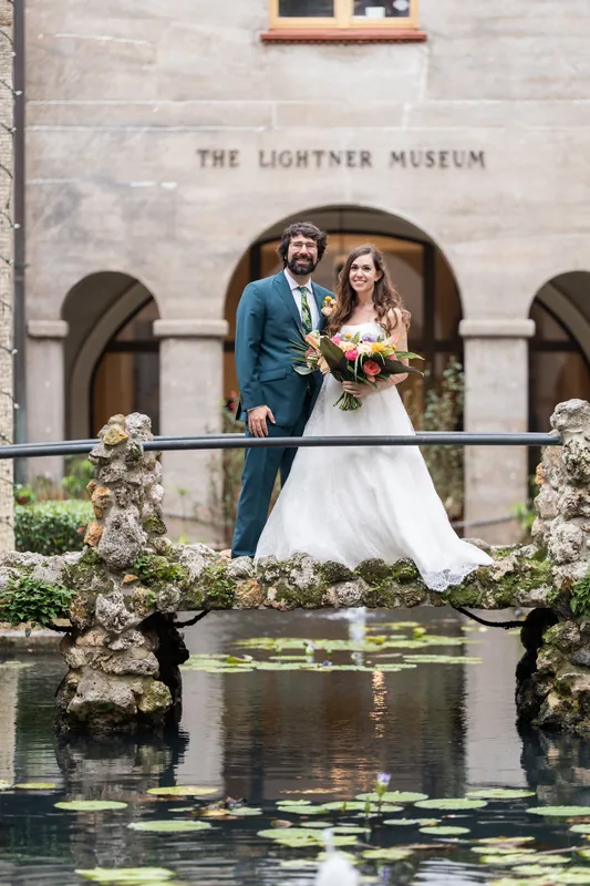 Two people in wedding attire stand on a stone bridge over a lily pond, white flowering shrubs flanking them at the Lightner Museum.