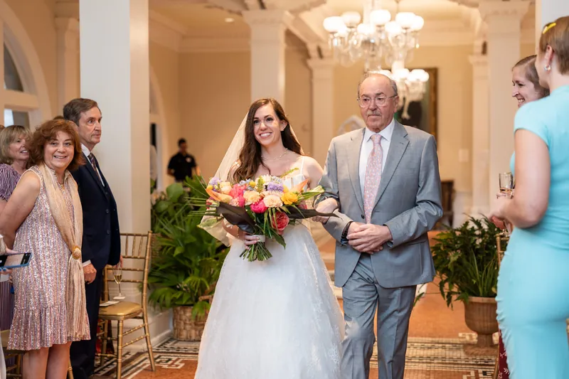 A person in a white gown walks through an arched hallway with a companion in a gray suit, guests and greenery visible around them.