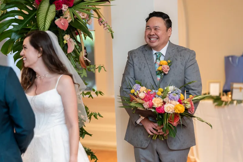 Someone in a gray suit holds a vibrant bouquet and smiles beside a person in a white sleeveless gown in a sunlit gallery.