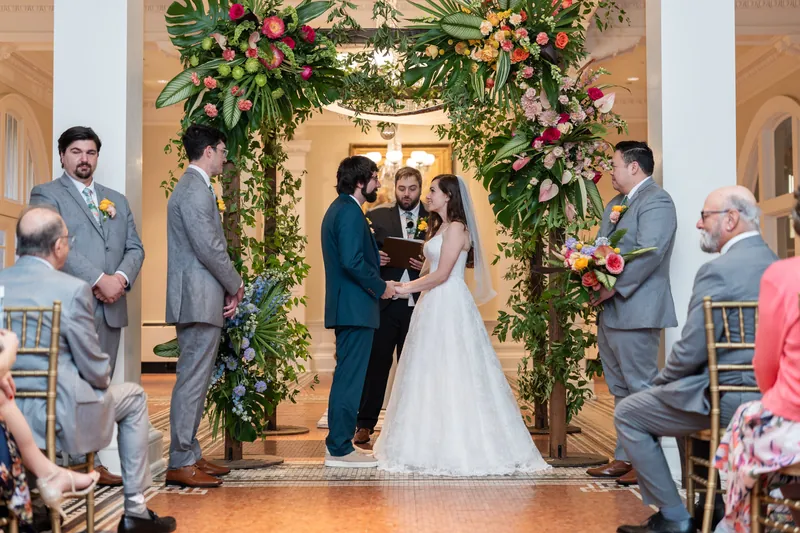 An officiant stands beneath a floral and greenery arch at the Lightner Museum, facing the couple during their ceremony as seated guests look on.