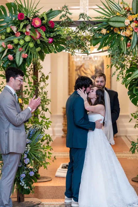 Two people kiss under a lush arch of roses and tropical foliage at the Lightner Museum as guests witness the moment.