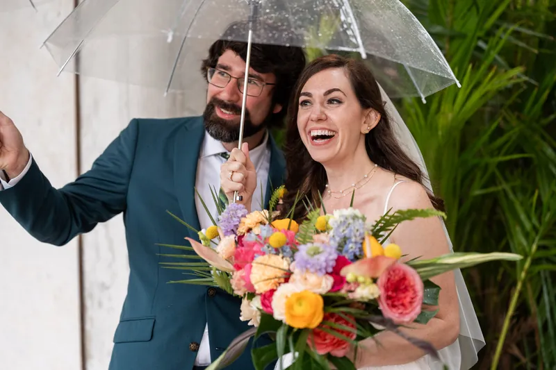 Two people laugh under a clear umbrella in the rain, one holding a vibrant bouquet of pink, purple, and yellow flowers.