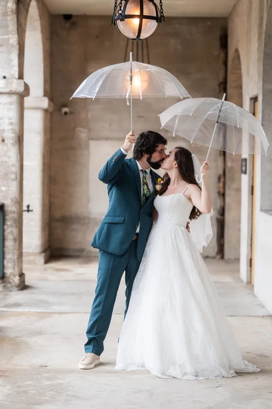 Jillian and Jason kiss under clear umbrellas in the Lightner Museum's columned corridor, both dressed in wedding attire.