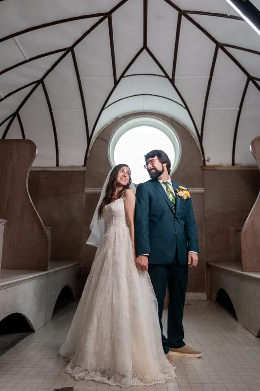 Jillian and Jason stand together beneath a geometric glass archway, she in a flowing white gown and he in a navy suit with a yellow boutonniere.