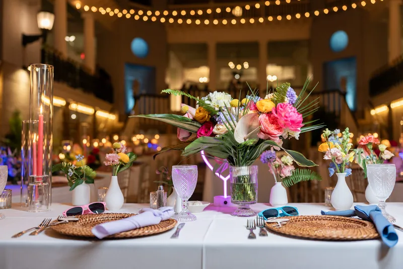 Vibrant pink and white floral centerpieces with greenery accent linen-draped reception tables under warm string lights at the Lightner Museum.