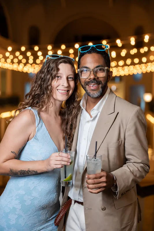 Two guests smile at the reception, the woman in a light blue dress and the man in a tan blazer, with glowing marquee lights visible behind them.