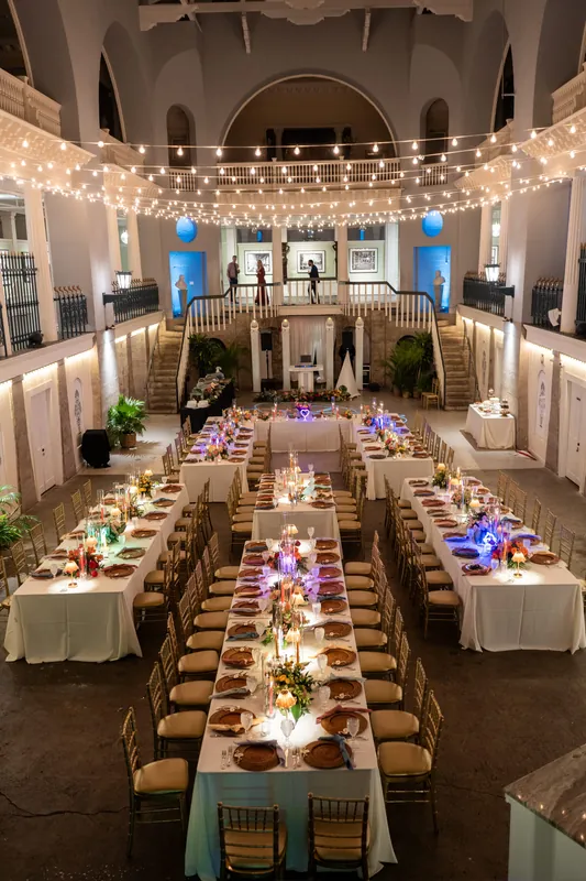 Elegant reception hall at Lightner Museum features long banquet tables with purple floral centerpieces, string lights overhead, and arched architectural details framing the space.