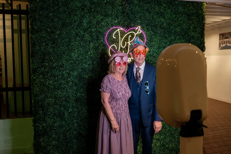 Two people in formal attire stand before a green hedge wall beneath a glowing neon monogram sign.