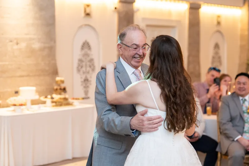 A man in a gray vest embraces a person in a white wedding dress while guests watch nearby.