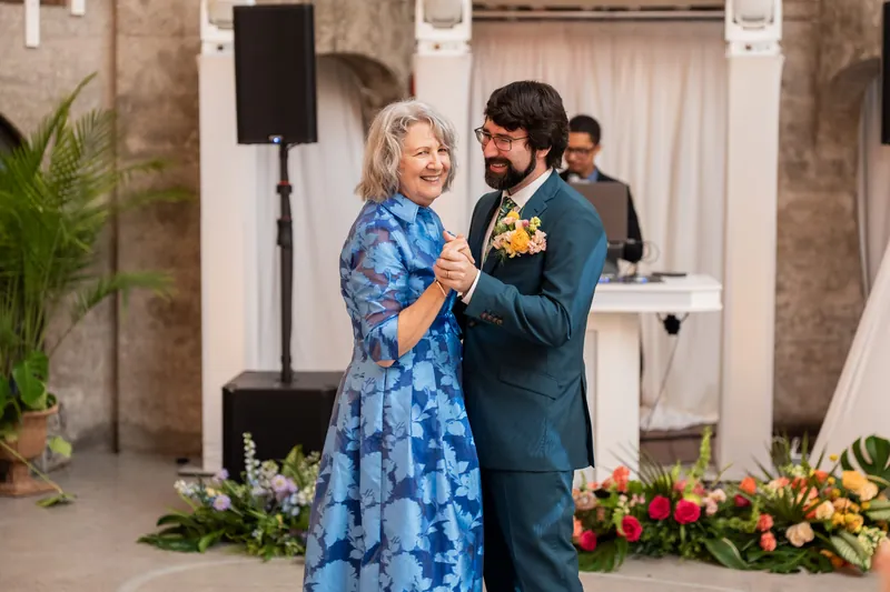 A woman in a blue patterned dress dances with a man in a teal suit as musicians and floral arrangements frame the moment.