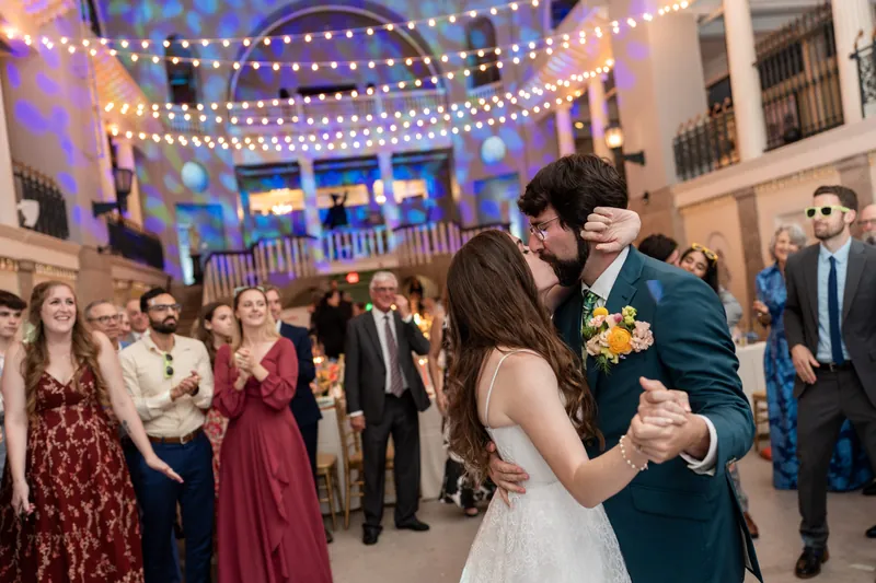 The couple dances under warm string lights while standing guests in formal wear look on during the reception.