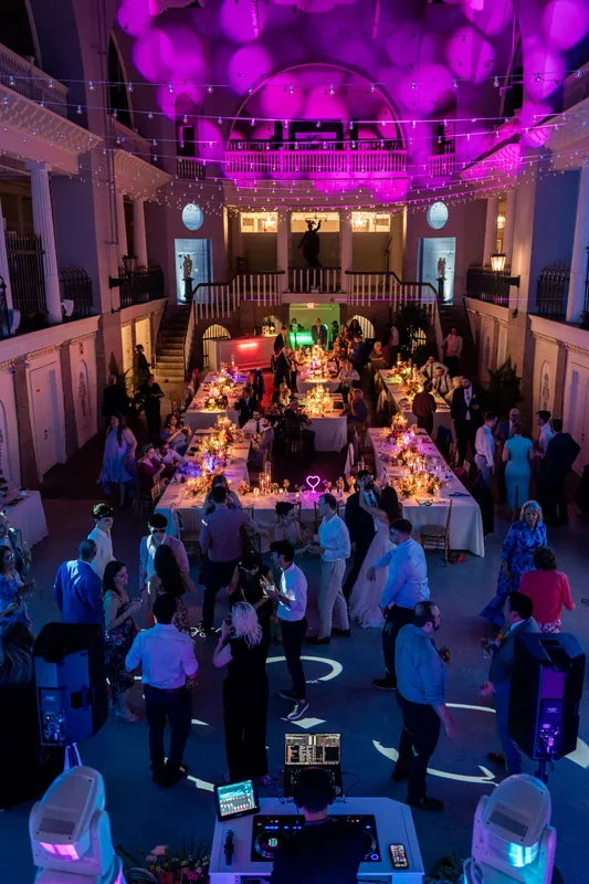 Overhead view of the Lightner Museum's grand hall filled with guests seated at long tables under magenta uplighting and a ornate ceiling.