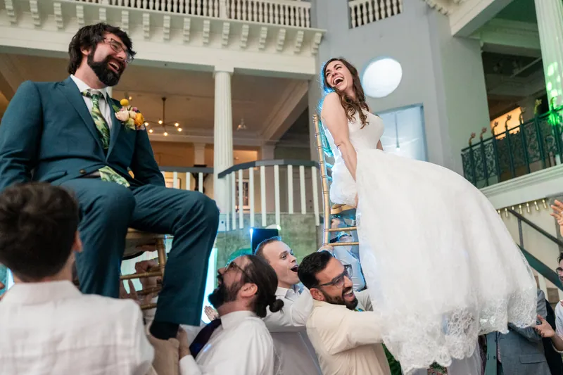 A person in a white gown sits on the shoulders of groomsmen while another person in a teal suit watches from a staircase at the Lightner Museum.