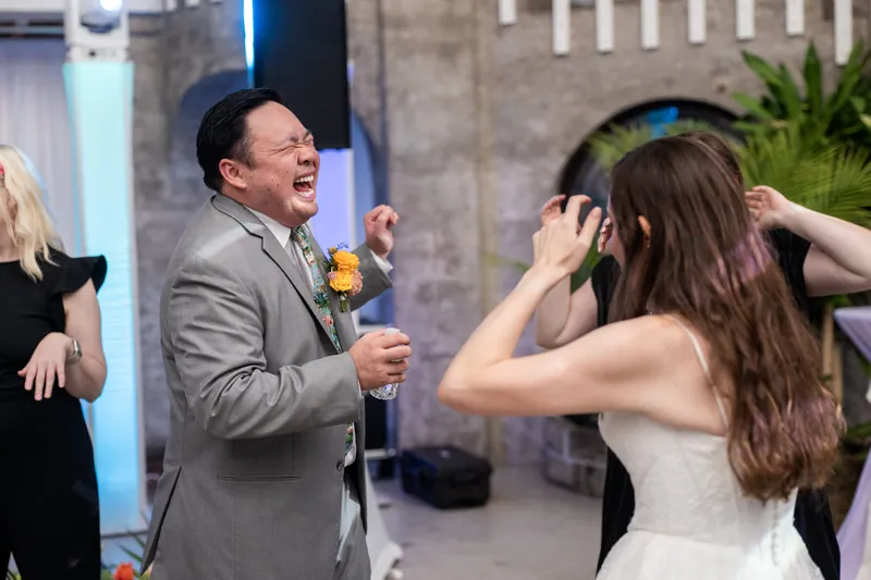 A man in a gray suit speaks into a microphone while a person in a white gown dances in front of him at the reception.