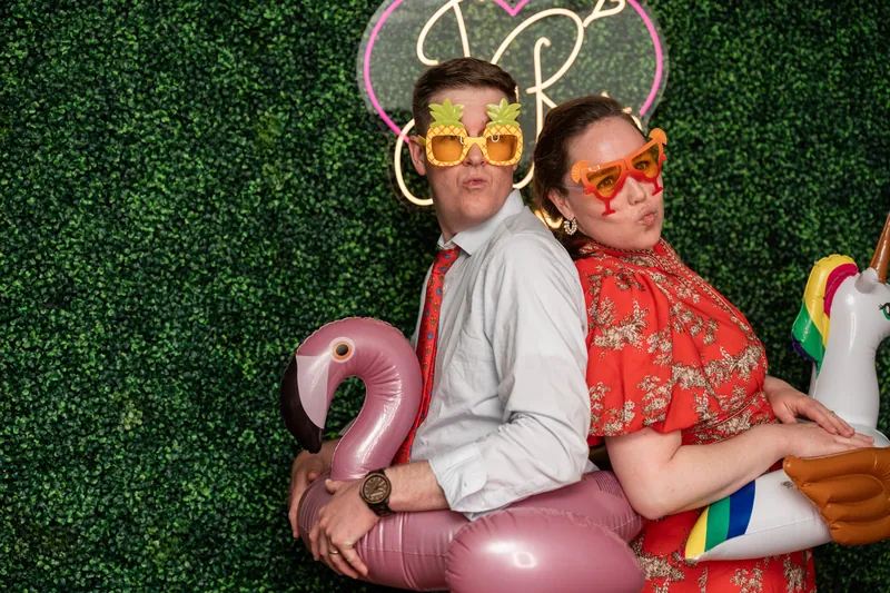 Two people pose back-to-back against a green hedge wall wearing novelty sunglasses and holding a pink number 2 balloon.