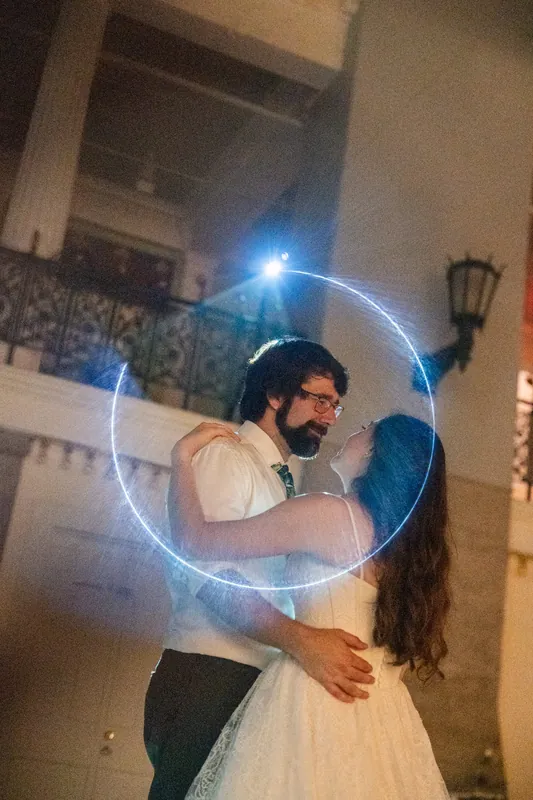 A couple embraces under a glowing blue circular light installation in an ornate interior hallway.