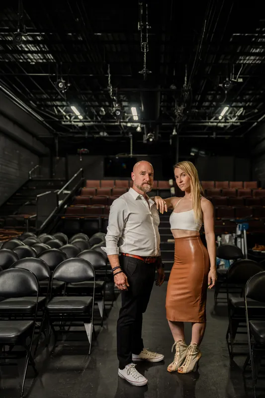 Man in gray button-up and woman in white crop top and copper skirt stand together on Limelight Theatre's stage, theater seats