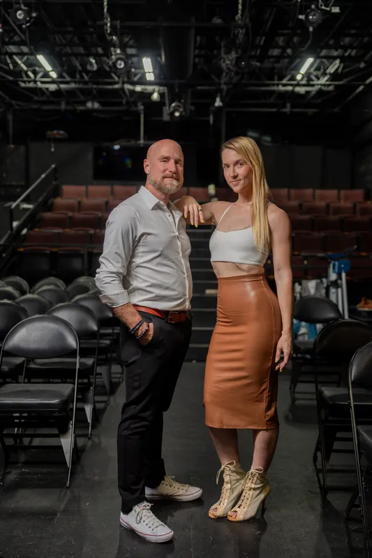 Man and woman stand close on Limelight Theatre's stage, the theater's brick wall and dark seating visible in the background.