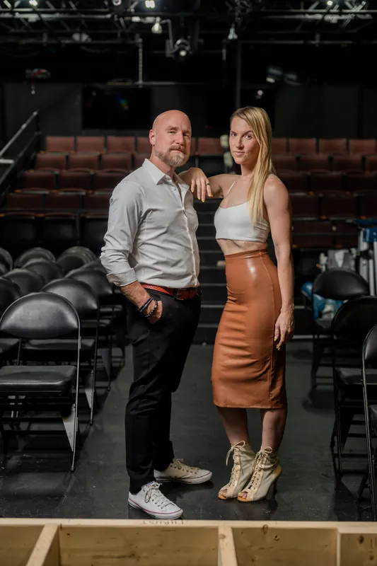 Man in gray shirt and woman in copper skirt embrace on Limelight Theatre's stage, theater seats and construction materials