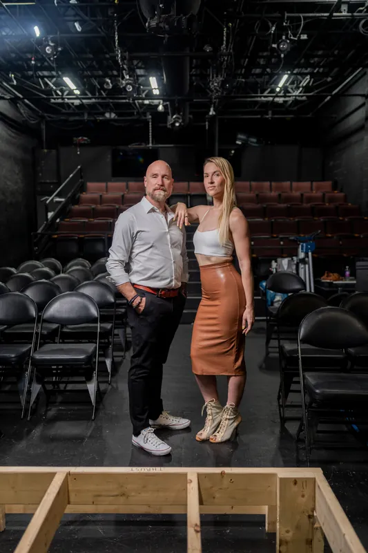 A man in a light blue shirt and woman in a tan skirt stand on the Limelight Theatre stage, with red theater seats