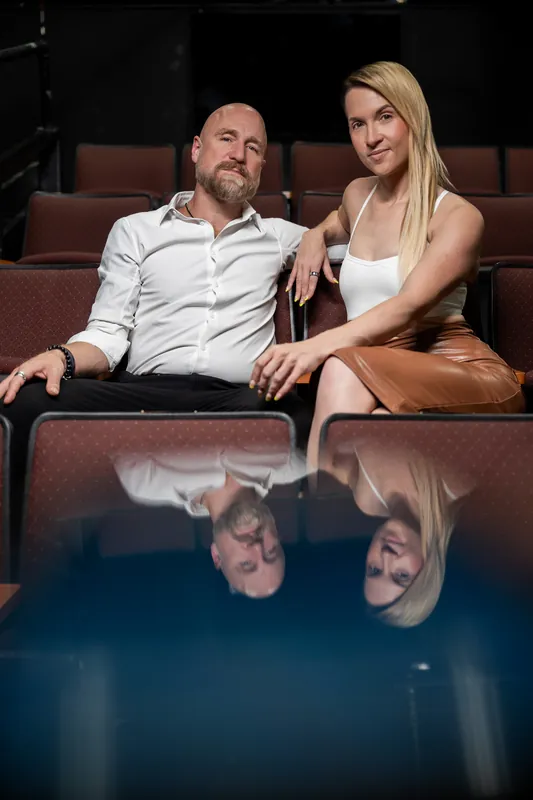 Seated in burgundy theater seats at Limelight Theatre, a bearded man in a white shirt and blonde woman in a white tank top