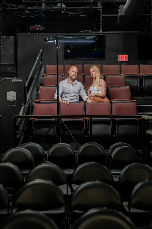 Framed by rows of empty black and burgundy seats, a man in a gray shirt and woman in a tan skirt sit together in Limelight