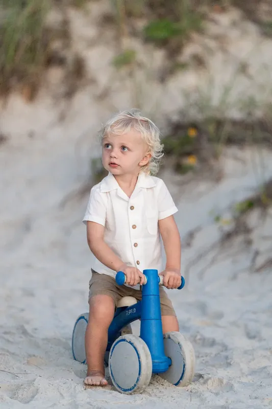 Toddler in white button-up shirt rides a blue tricycle on the sand at St. Augustine Beach.