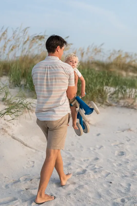 Adult carries a young child in blue shorts while walking barefoot across the sand at St. Augustine Beach.