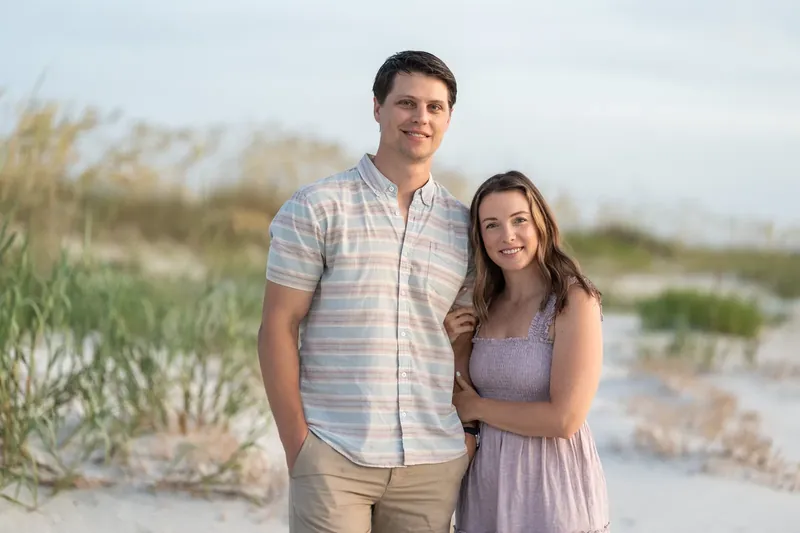 Jourdan smiles while standing arm-in-arm with partner on the beach, dunes visible in the background.
