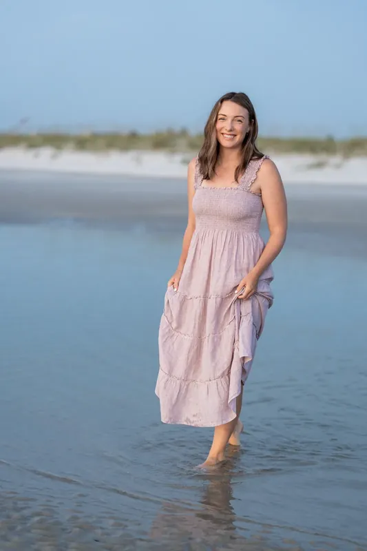 Jourdan walks through the shallow water in a lavender dress, smiling toward the camera.