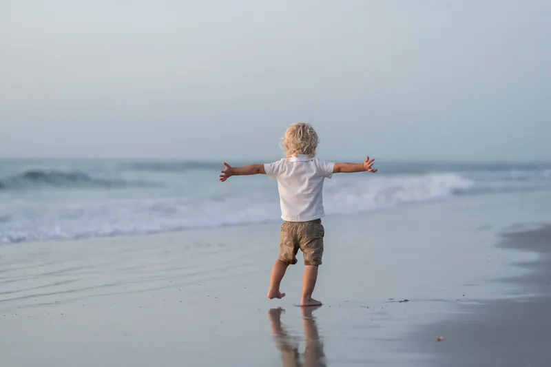 Young child stands arms outstretched in shallow surf at St. Augustine Beach, wearing white shirt and tan shorts.