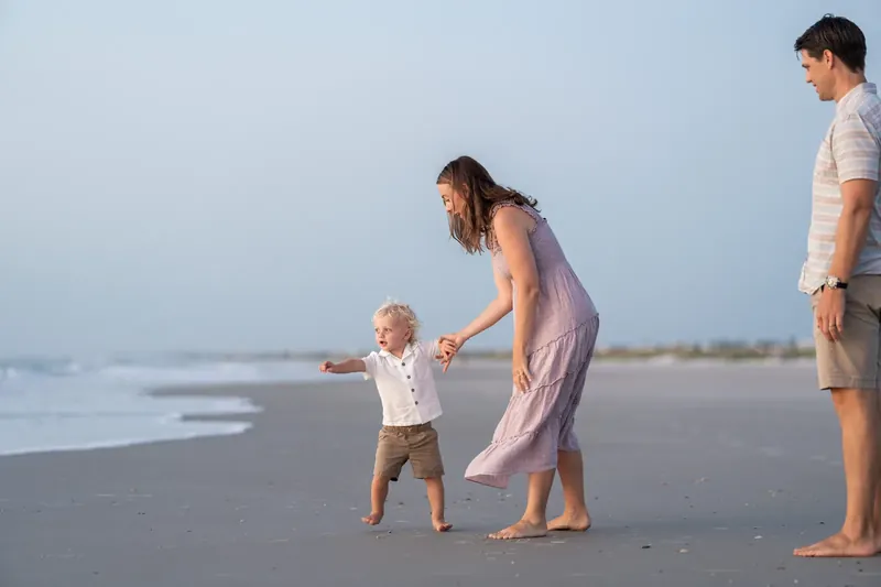 Adult in pink dress holds toddler's hands while walking on wet sand as another adult watches nearby.