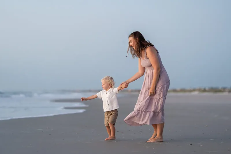 Adult in mauve dress guides toddler across the beach, both facing the ocean with calm waters behind them.