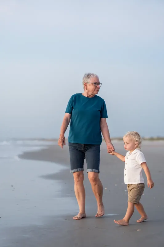 Adult in blue shirt and shorts holds toddler's hand while strolling along the shoreline at St. Augustine Beach.
