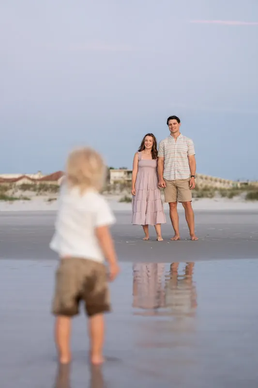 Young child runs toward Jourdan and partner standing together on the wet sand at St. Augustine Beach at dusk.