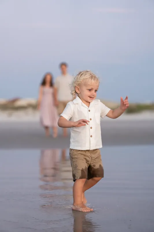 Blonde toddler splashes through shallow water while Jourdan and partner watch from behind on the beach.