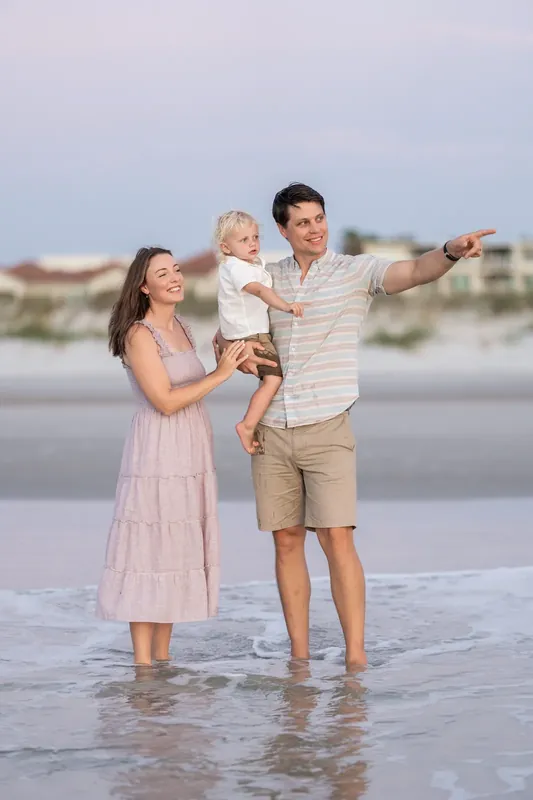 Jourdan holds their child while partner points toward the horizon, both standing in the shallow surf.