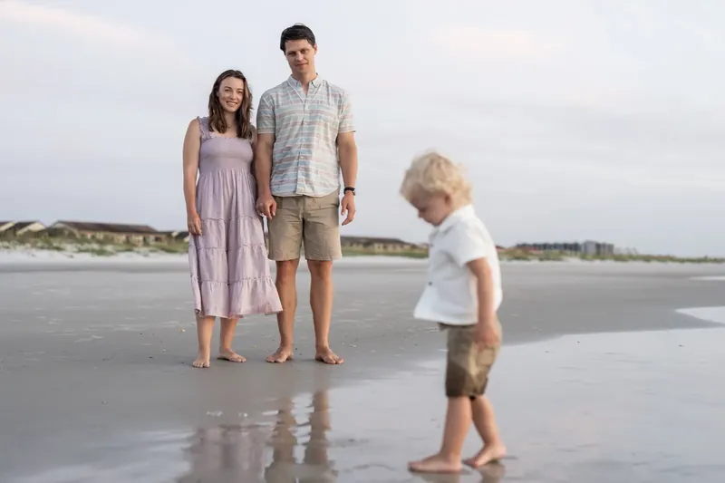 Partner and Jourdan stand together on the beach while their child runs past them on the wet sand.