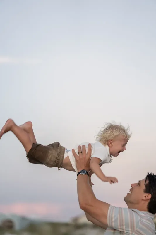An adult lifts a laughing blonde toddler horizontal above their shoulders against the beach backdrop.