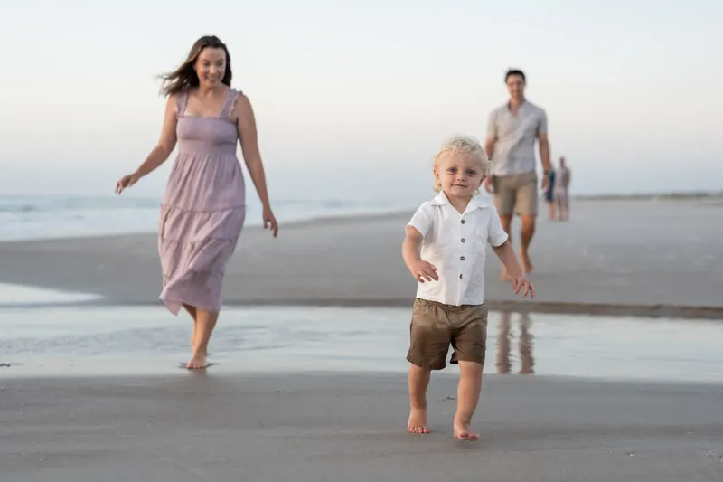 A woman in a mauve dress walks barefoot on the sand as a young child in a white shirt races ahead toward the water.