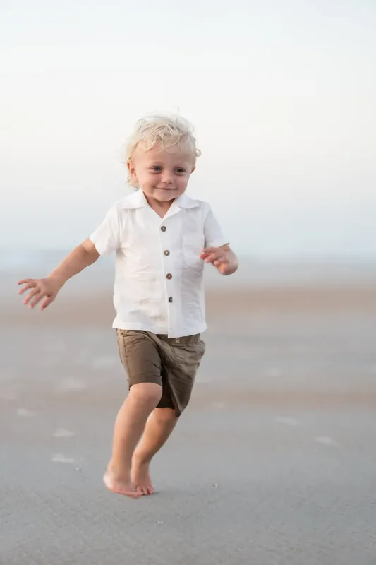A blonde toddler in a white button-up shirt and brown shorts runs barefoot across the wet sand at the shoreline.