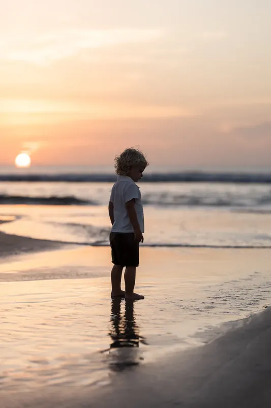 Child in white shirt stands alone at the water's edge at sunset, gazing toward the horizon at St. Augustine Beach.