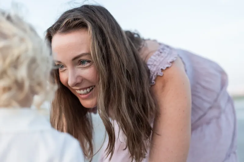 Woman with long dark hair smiles brightly at the camera, wearing a lavender top with lace details.