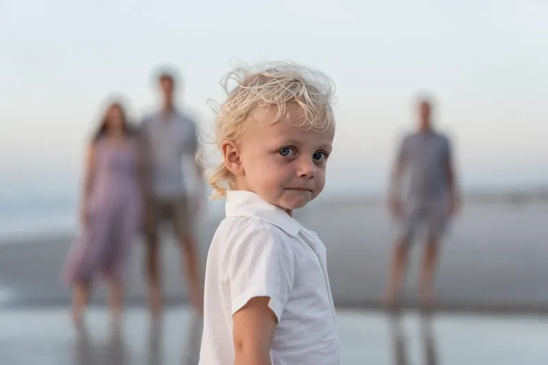 Blonde toddler in white shirt looks back toward the camera as family members stand together in the shallow water behind.