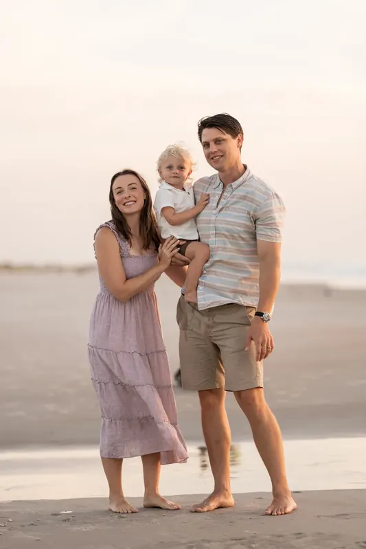 Jourdan and their partner hold a young child between them while standing barefoot in the sand at St. Augustine Beach.